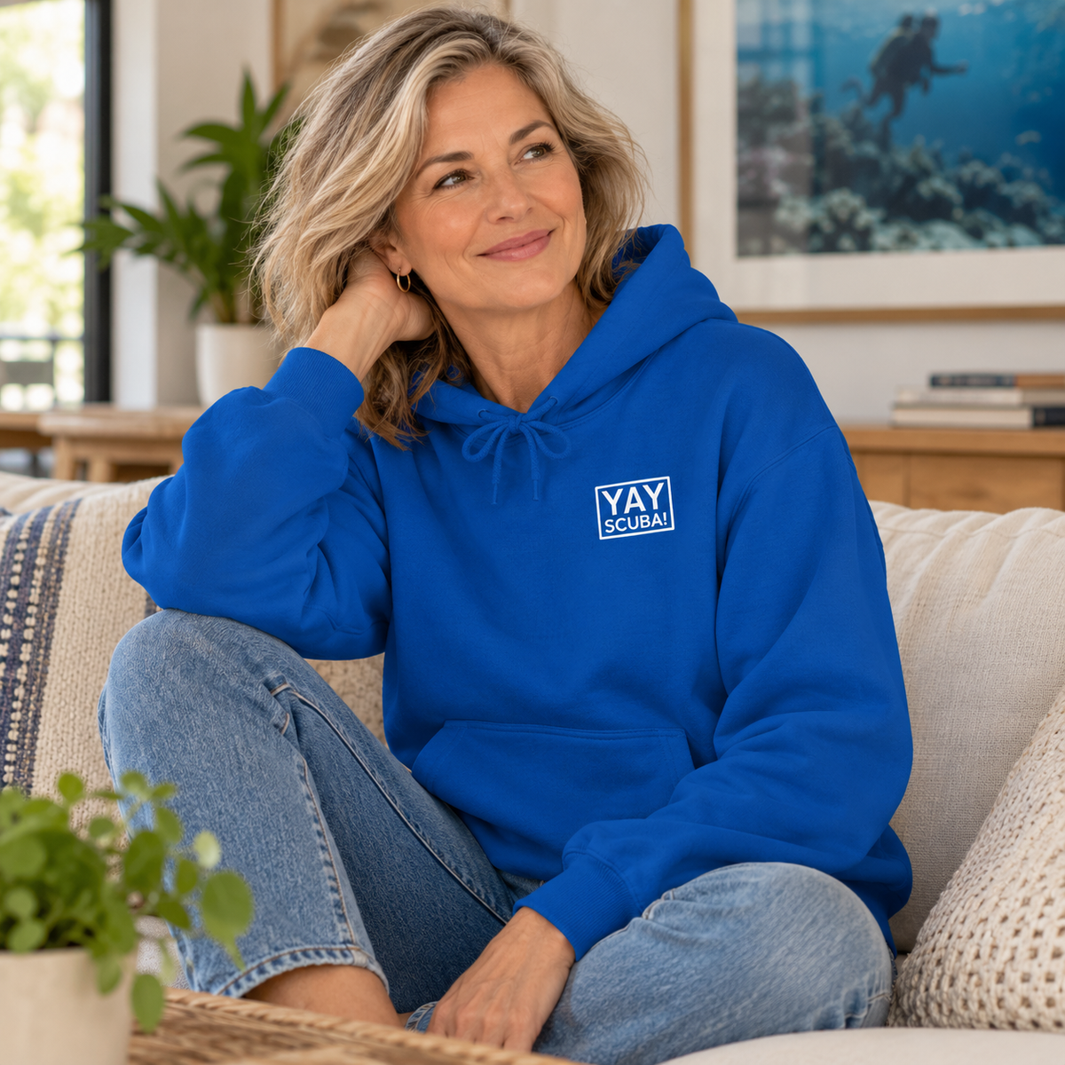 A woman sitting on a beige couch wearing a bright blue hoodie with a white 'YAY SCUBA' logo on the chest, paired with blue jeans in a cozy living room setting.