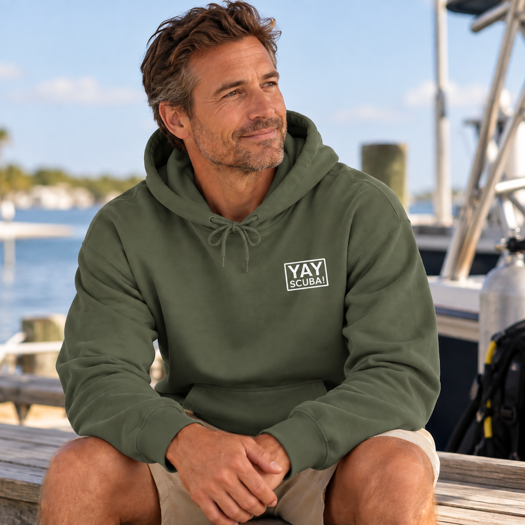 A man sitting on a wooden dock wearing an olive green hoodie with a white 'YAY SCUBA' logo on the chest, with water and boats in the background.