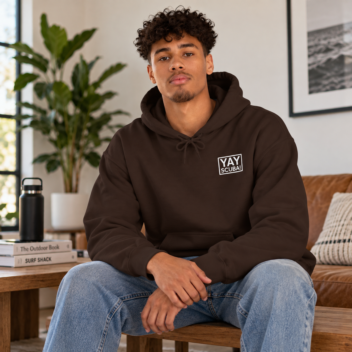 A man wearing a dark brown pullover hoodie with a white 'YAY SCUBA' logo on the left chest, sitting on a wooden bench in a modern living room setting.