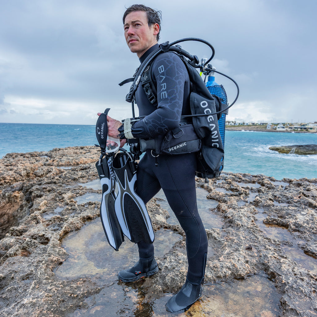 A man wearing a Oceanic Photon Pro Men's BCD stands on rocky coastal ground with the ocean and cloudy sky in the background, holding a pair of black-and-white flippers and carrying an oxygen tank.