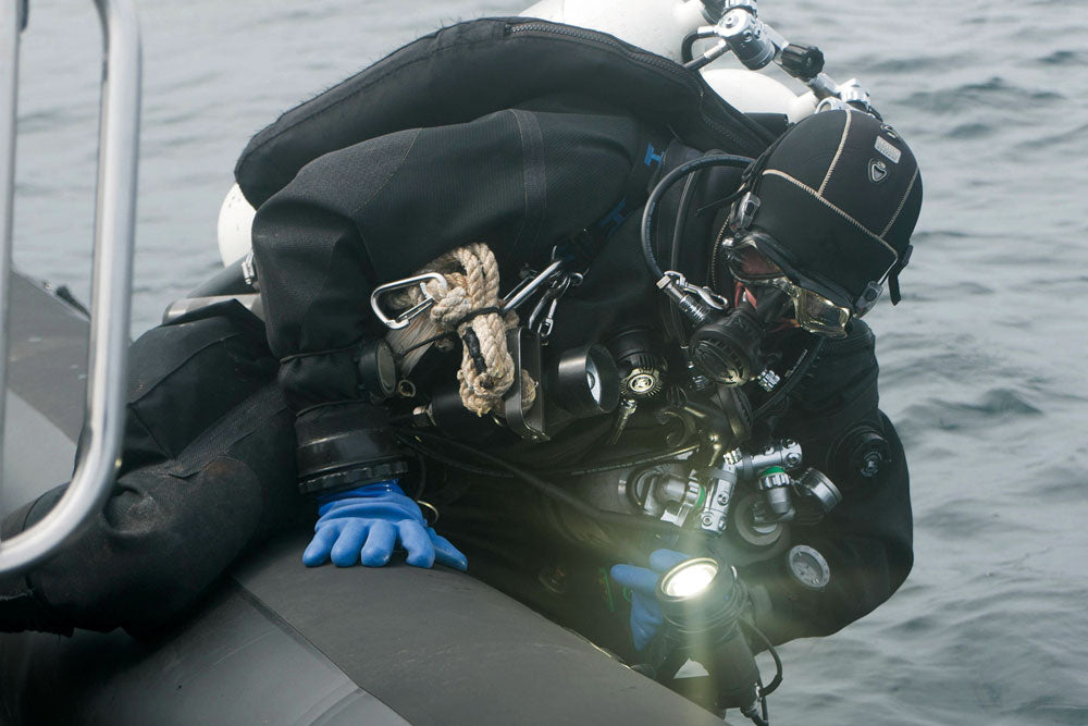 A person in full black scuba diving gear with a helmet, mask, tank, and blue SI TECH Quick Glove leans over the side of a small boat in open water, holding a bright dive light.