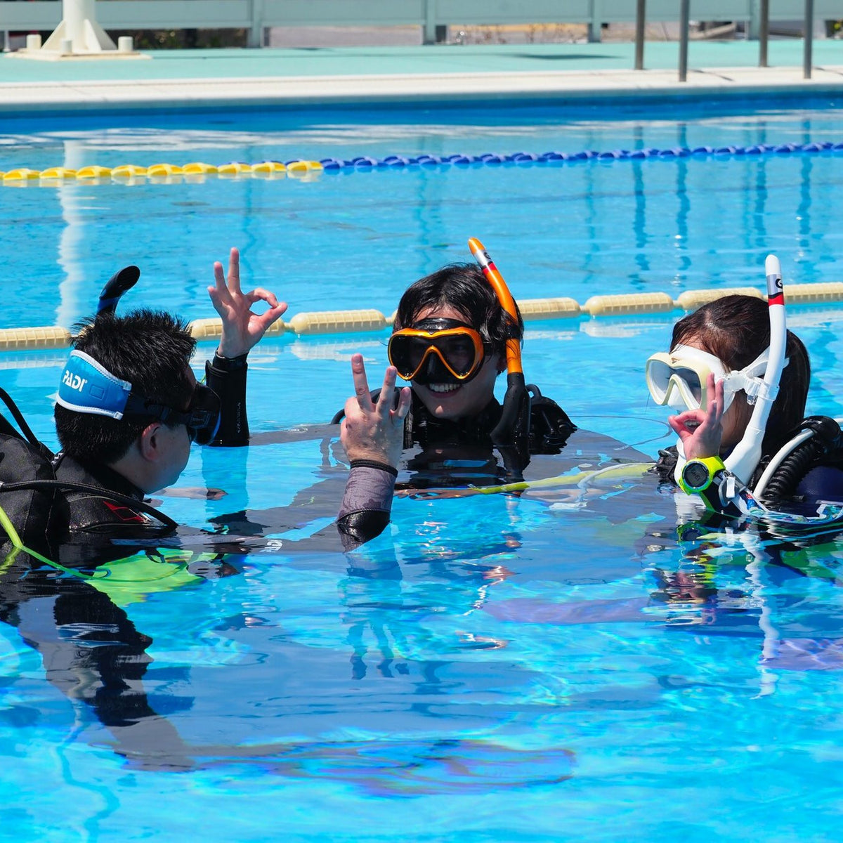 Three people in a swimming pool wearing scuba diving and snorkeling gear, including masks, snorkels, regulators, and wetsuits, with yellow and blue lane lines in the background.